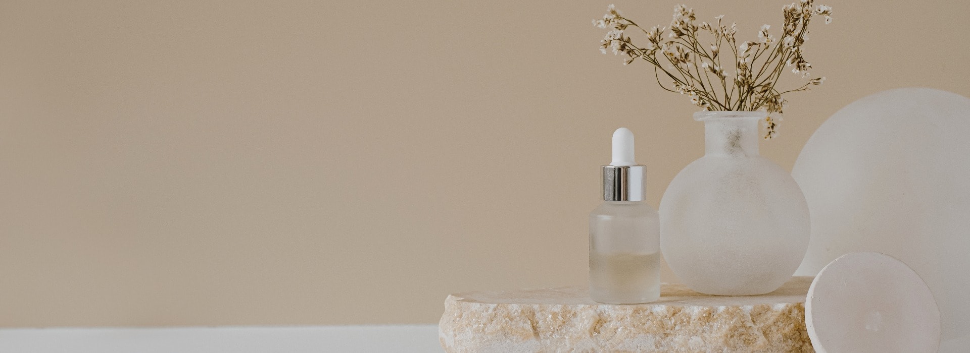 Aesthetic minimalist beauty care therapy concept. Organic serum oil cosmetics bottle on stone with flowers against neutral beige background. Body skin, face treatment product composition